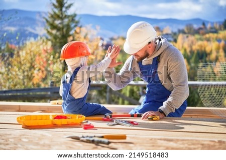 Father with toddler son building wooden frame house. Male builder giving high five to kid on construction site, wearing helmet and blue overalls on sunny day. Carpentry and family concept.