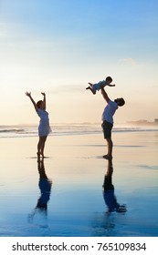 Father Tossing High In Air Baby Son, Mother Jumping By Water Pool. Happy Family Walk With Fun By Sunset Black Sand Beach With Sea Surf. Active Parents, Outdoor Activity On Summer Vacation With Kids.
