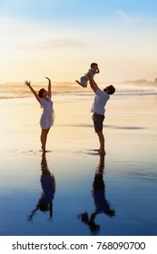 Father Tossing High In Air Baby Son, Mother Jumping By Water Pool. Happy Family Walk With Fun By Sunset Black Sand Beach With Sea Surf. Active Parents, Outdoor Activity On Summer Vacation With Kids.