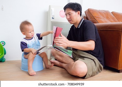 Father Training His Son to Use Potty, Cute little Asian 1 year old / 18 months toddler baby boy child sitting on potty and reading book with young dad in living room at home. Potty Training concept.