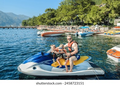 Father with two small sons wearing life jackets is seated on personal watercraft near beach with boats and pine trees in a seaside resort - Powered by Shutterstock