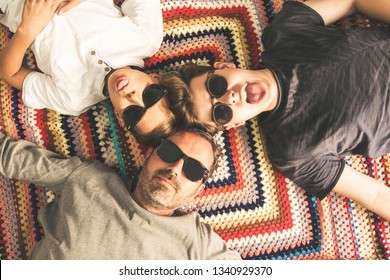 Father And Two Sons Enjoying Together Lying On A Colorful Blanket. Three Men Of Different Ages With Black Sunglasses. Top View Of A Couple Of Teen Making Tongue And Their Dad.