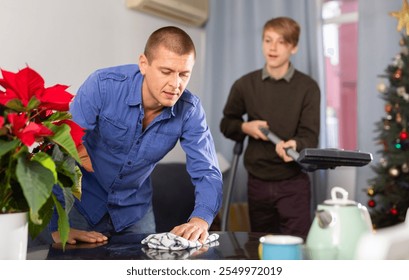 Father wipes the table, son vacuums the room before celebrating the new year - Powered by Shutterstock