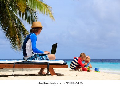 Father Working On Laptop While Kids Play With Sand On Beach