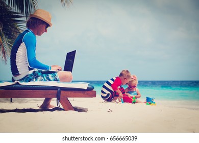 Father Working On Laptop While Kids Play At Beach