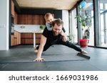 Father working out, doing single arm plank with his jolly infant baby riding on his neck. At home apartment.