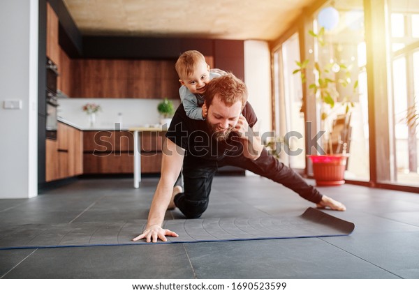 Father\
working out, doing single arm plank with his jolly infant baby\
riding on his neck. Stay at home apartment. Family quarantine,\
domestic life in self-isolation. Sunset light\
