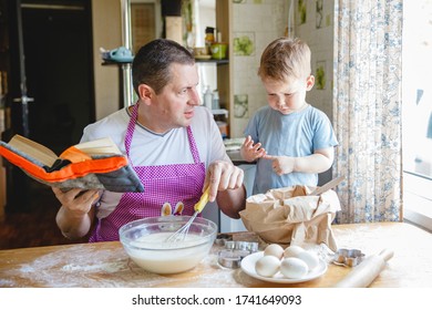 A Father With A Young Son At The Kitchen Table At Home Preparing Dough. Dad Is Reading A Book, And The Boy Is Busy In The Flour.