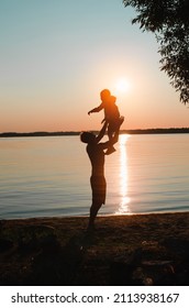 Fatherhood, Childhood, Family Vacation. Silhouettes Of Father And Son Having Fun Together On Beach At Sunset, Outdoors. Side View Of Dad Holding Child In Arms, Throwing Up Air Against Sky.