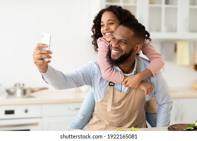 Fatherhood Concept. Portrait of cheerful african american father taking selfie with his little daughter while cooking in the kitchen together at home, giving her piggyback ride, using smartphone - Powered by Shutterstock