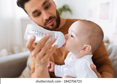 Fatherhood. Young Father Giving His Little Baby Toddler Bottle Of Water Feeding Him Sitting On Sofa At Home. Paternity Leave, Child Care, Parenting And Parenthood Concept. Selective Focus