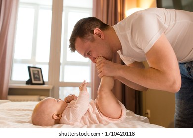 Fatherly Love. Dad Kissing The Legs Of A Baby Lying On The Bed