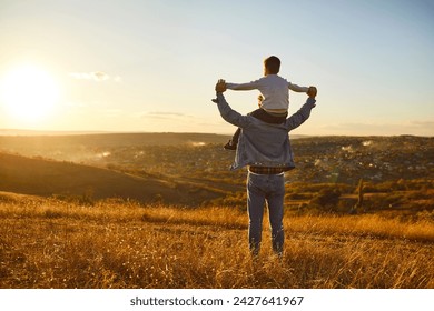 Fathers day. Back view of a little child boy sitting on his fathers shoulders holding hands and looking into the distance enjoying sunset. Father walking with son outdoors. - Powered by Shutterstock