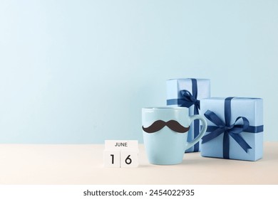 Father's Day celebration concept featuring a blue mug with a whimsical mustache design, neatly wrapped gifts, and a calendar marking June 16, all set against a soft blue backdrop - Powered by Shutterstock