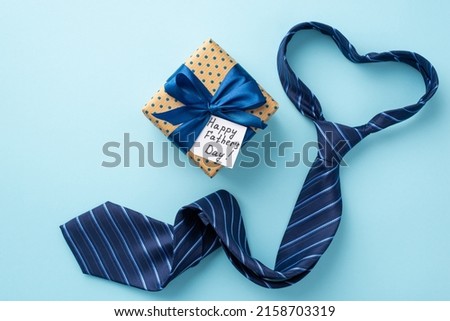 Father's Day concept. Top view photo of heart shaped blue necktie polka dot giftbox with satin ribbon bow and postcard on isolated pastel blue background