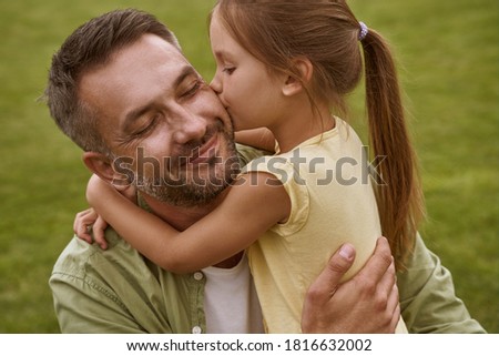 Fathers day. Cute little girl hugging and kissing her happy dad while sitting on a green grass