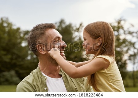 Fathers day. Cute little girl playing with her daddy while visiting park on a summer day, touching his cheeks and smiling