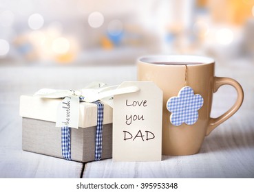 Father's Day Holiday Greeting Card.Mug Tea Coffee And Present Gift Box Tag On Wooden Table Blur Lights Background Empty Space.Love You Dad Message.