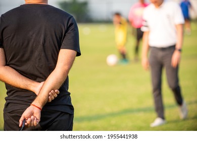 Fathers Standing And Watching Their Sons Playing Football In A School Tournament On A Sideline With A Sunny Day. Sport, Outdoor Active, Lifestyle, Happy Family And Soccer Mom And Soccer Dad Concepts.