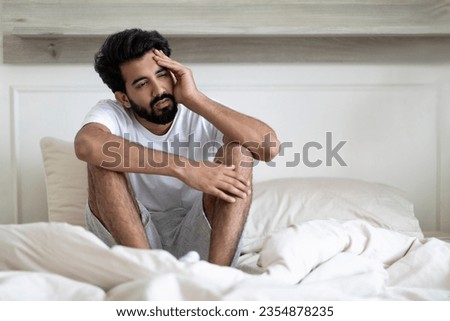 Fatigue Concept. Tired Young Indian Man Sitting In Bed At Home, Exhausted Upset Millennial Eastern Guy Waking Up In Bedroom, Feeling Unwell, Having Headache Or Suffering Insomnia, Free Space
