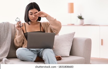 Fatigue From Work. Tired Sleepy Asian Lady Feeling Eyestrain, Holding Glasses, Rubbing Dry Irritated Eyes, Working On Laptop, Sitting On The Couch In Living Room. Woman Touching Hurt Sore Eye