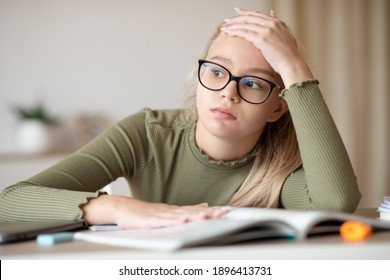 Fatigued blonde teen girl with glasses sitting at home with book, doing homework, touching her forehead and looking at copy space. Bored schooler got tired of studying, missing her friends