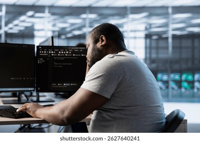 Fatigued engineer falling asleep while typing on computer in server hub, monitoring data center energy consumption. Sleepy employee struggling to stay awake while inspecting rackmounts