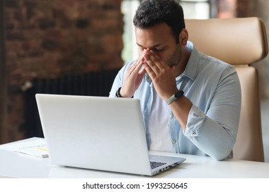 Fatigued Indian Man Using Laptop For Working, A Guy In Casual Wear Is Touching And Massaging The Bridge Of The Nose, Feels Tired And Burnout, Sitting At The Desk In Modern Office With Eyes Closed