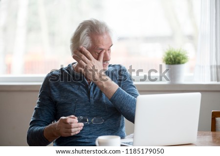 Fatigued mature old man taking off glasses suffering from tired dry irritated eyes after long computer use, senior middle aged male feels eye strain problem or blurry vision working on laptop at home