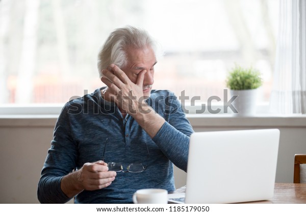 Fatigued mature old man taking off glasses suffering\
from tired dry irritated eyes after long computer use, senior\
middle aged male feels eye strain problem or blurry vision working\
on laptop at home