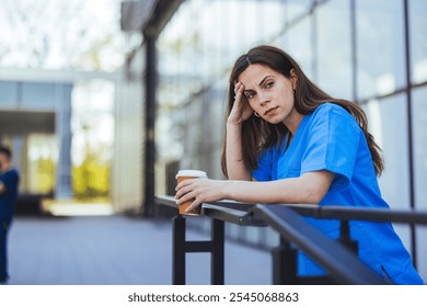 A fatigued nurse in blue scrubs leans on a railing, holding a coffee cup, reflecting exhaustion. The outdoor hospital setting underlines healthcare challenges and the need for moments of respite. - Powered by Shutterstock