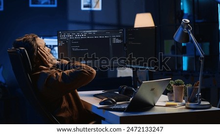 Fatigued software programmer working overnight at home, taking break from coding to stretch at desk chair. Overworked IT professional feeling exhausted, pausing tasks to rest, camera A