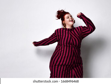 Fatty ginger girl with bun hairstyle, in black and purple striped dress and golden earrings. She is smiling, dancing and having fun while posing isolated on white background. Close up, copy space.