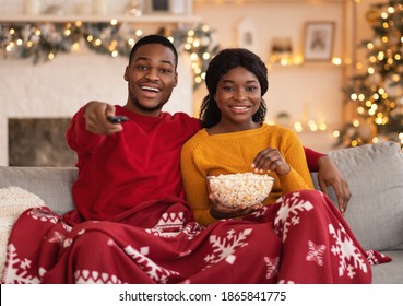 Favorite movie together at home and self-isolation during lockdown. Smiling young african american lady with popcorn, guy with remote control, covered with blanket watch film in interior with garlands