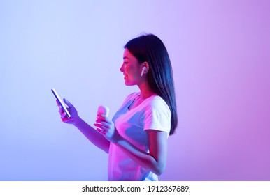 Favorite Music For Relax And Sports During Covid-19. Smiling Beautiful Asian Teen Lady In Wireless Headphones Looks In Smartphone And Holds Box For Headset, In Neon, Studio Shot, Side View, Free Space