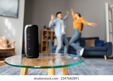 Favorite Music. Unrecognizable Asian Couple Listening To Song Via Wireless Smart Speaker Having Fun, Jumping And Dancing Together At Modern Home Interior. Selective Focus On Musical Column