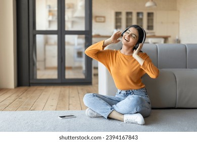 Favorite Song. Smiling Young Indian Woman Wearing Wireless Headphones Listening Music On Smartphone While Relaxing At Home, Beautiful Eastern Female Sitting On Floor With Closed Eyes, Free Space - Powered by Shutterstock