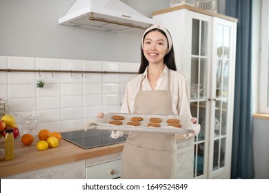 Favourite buisness. Young pretty woman standing at home in the kitchen with a baking sheet in her hands, smiling. - Powered by Shutterstock