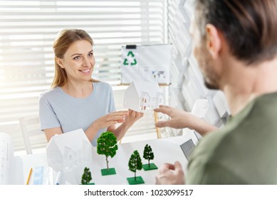 Favourite Job. Beautiful Happy Fair-haired Young Woman Smiling And Working With Her Colleague On An Eco-friendly Project And Holding A House Miniature While Sitting At The Table