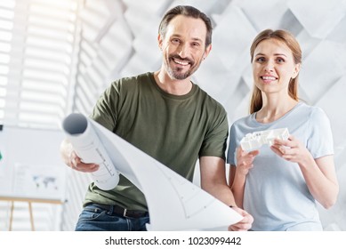 Favourite Job. Handsome Delighted Bearded Man Smiling And Holding A Drawing And Working On The Project With His Colleague And She Holding A House Miniature