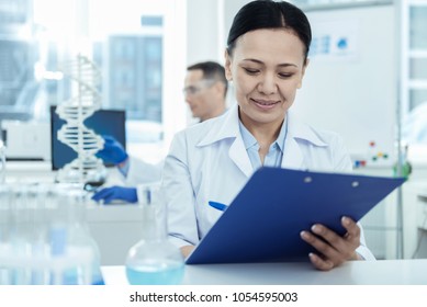 Favourite Work. Beautiful Joyful Dark-haired Researcher Smiling And Wearing A Uniform While Taking Notes And Her Colleague Sitting In The Background