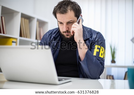 FBI agent working in his office talks on mobile an looking at laptop