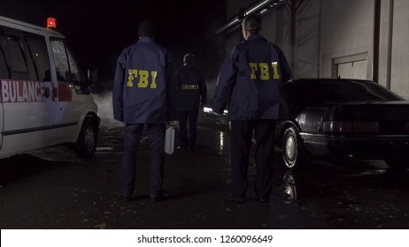 FBI agents work at the scene at night, police car with lights and ambulance background. Back view on three FBI agents go towards criminal scene