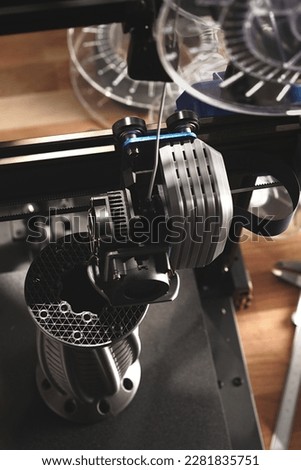 FDM 3D printer produces complex flange part from silver grey material on wooden desk. vertical orientation. view on print head, infill structure. selective focus. additive manufacturing concept