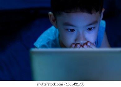 Fear or excite face of kid. Little boy age 6 years old use 2 hands close his mouth when seeing violent or sensitive content on tablet with dark lowlight in bedroom and blue light reflection on facial.