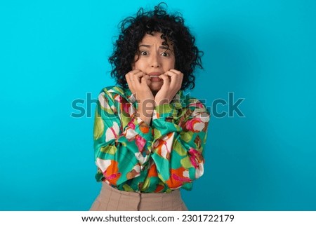 Fearful young arab woman wearing colorful shirt over blue background keeps hands near mouth, feels frightened and scared,  has a phobia,  Shock and frighted concept.