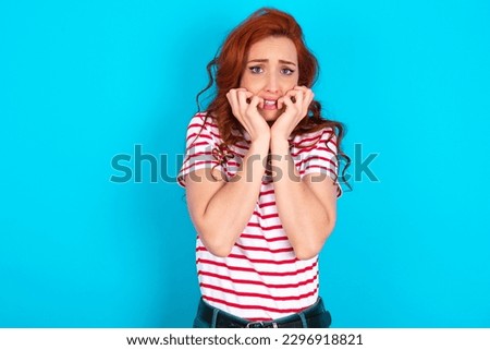 Fearful young redhead woman wearing striped T-shirt over blue background keeps hands near mouth, feels frightened and scared,  has a phobia,  Shock and frighted concept.