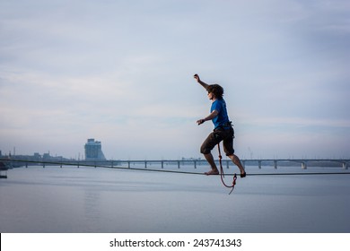 Fearless Highliner Walking On Tight Rope Over The River In Front Of The Bridge And Building With National Ukrainian Symbol In Dnipropetrovsk, Ukraine