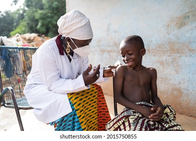 Fearless little African boy watching at the needle of a vaccine-filled syringe to be injected into his arm by a gentle black nurse
