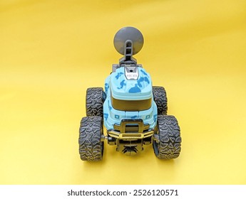 features a blue and gray toy monster truck with large, rugged wheels and a satellite dish mounted on top. The truck has a camouflage pattern and is positioned against a bright yellow background.